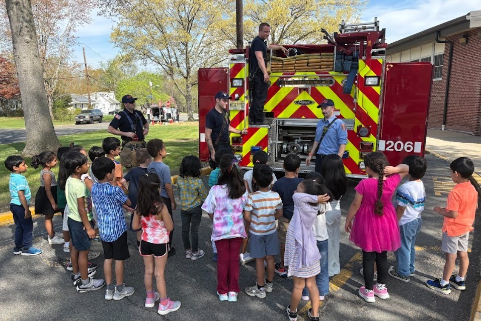 A group of children stands in a circle facing a fire truck with firefighters on it.