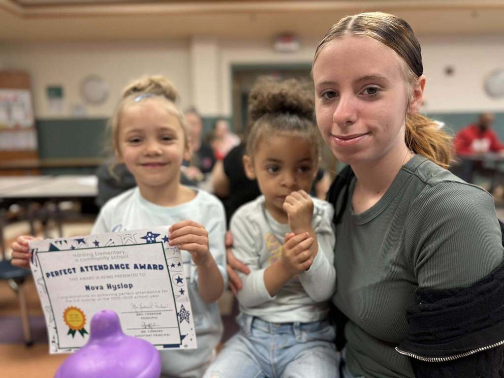 A student holding her perfect attendance award and sitting next to women and another child. 