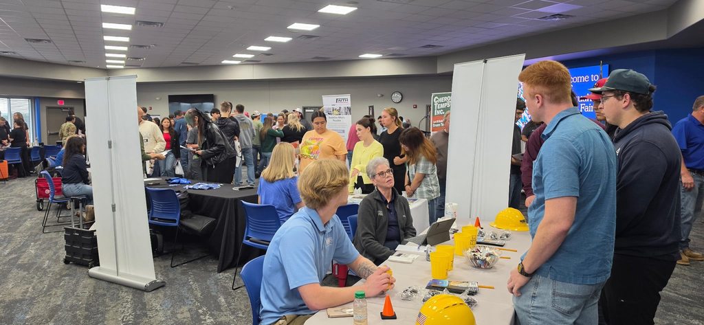 Group of people in a hall, some wearing yellow hard hats, standing and seated at tables with various items.