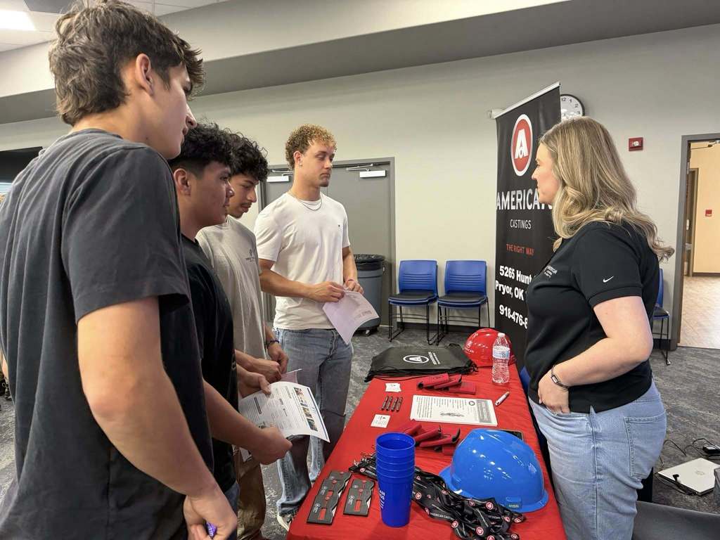 Five people gather around a table with helmets and tools, listening to a woman in a black shirt.