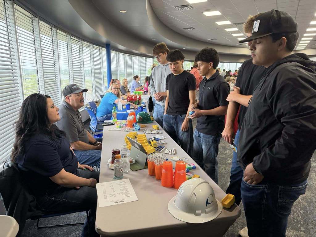 Several people stand and sit at a table with items on it in a room with large windows.