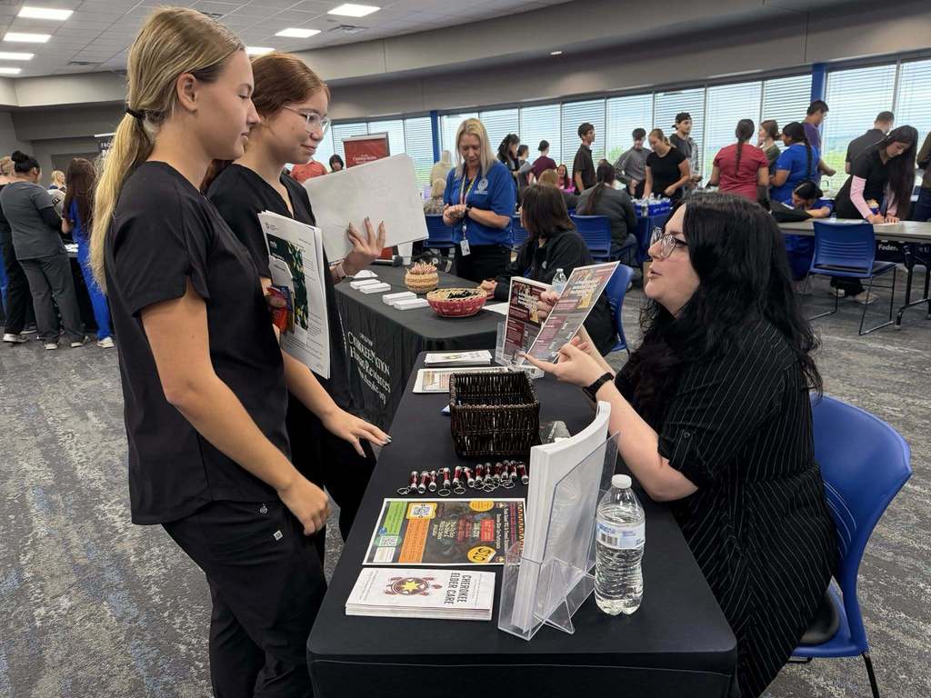 People are gathered in a room. A woman sits at a table, reading materials while others stand behind her.