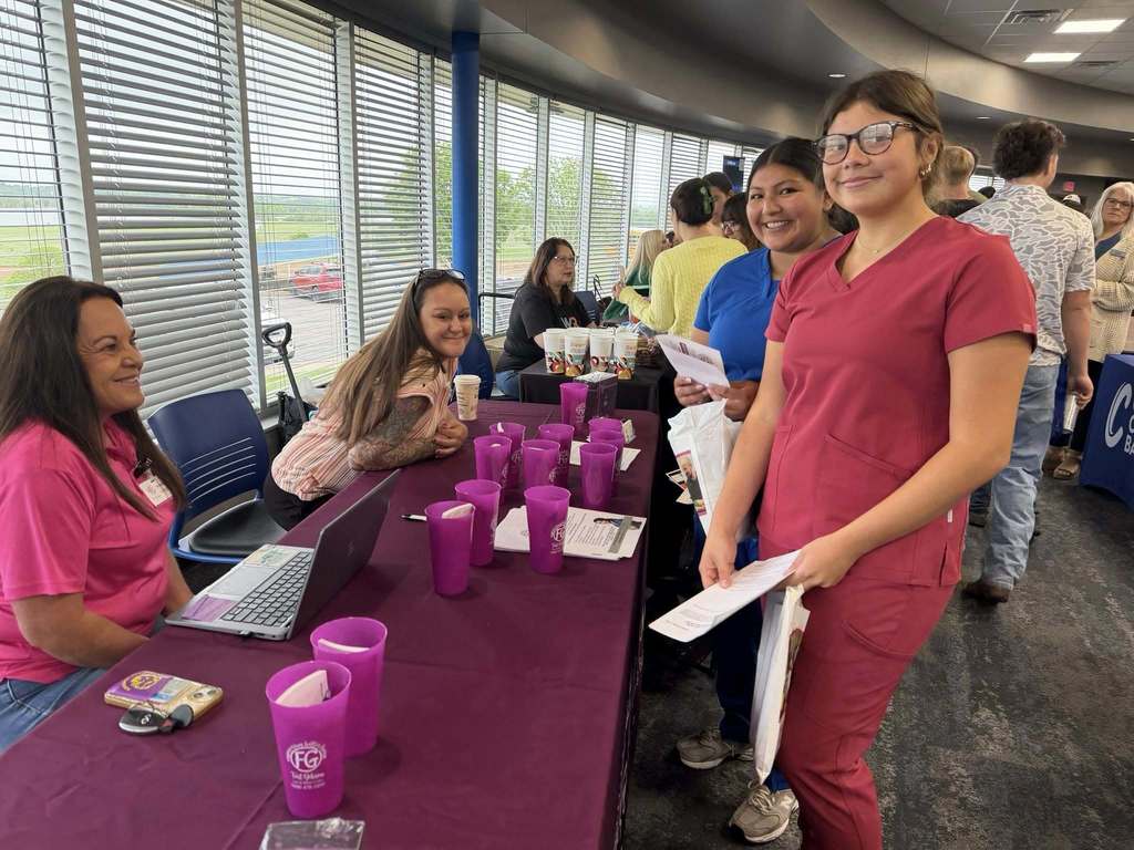 A group of people, some in uniforms, seated and standing in a room with a table featuring purple cups, papers, and a laptop.