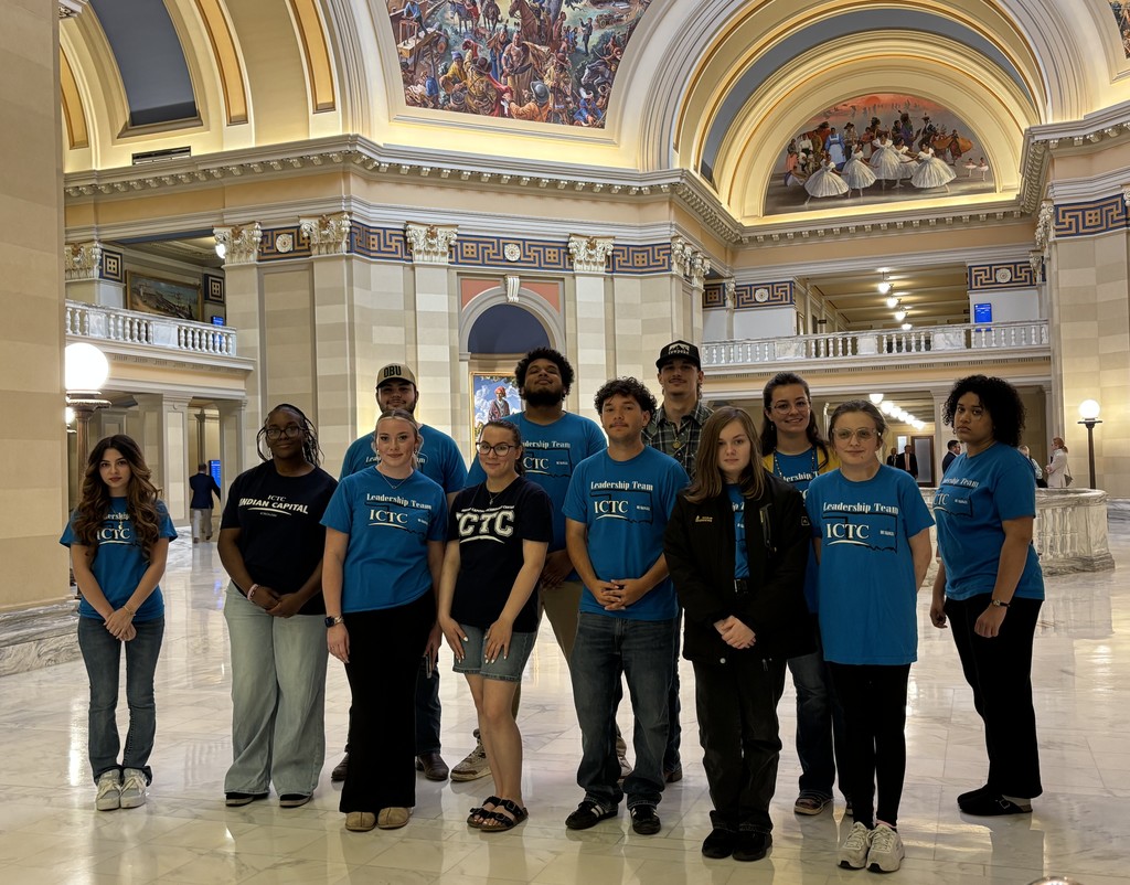 A group of people wearing blue shirts with various text on them stand in a large, ornate hall with high ceilings.