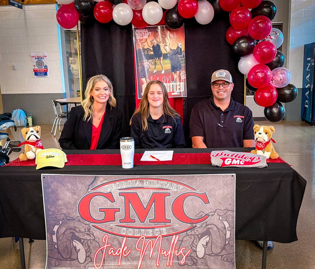 Three people seated behind a table with a GMC banner. Two women and one man with red balloons in the background.