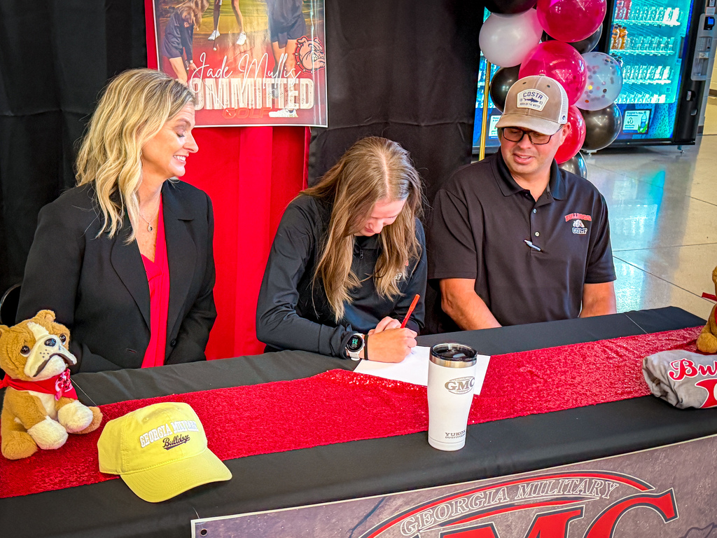 Three people, two women and a man, seated at a table with a red banner, signing documents.