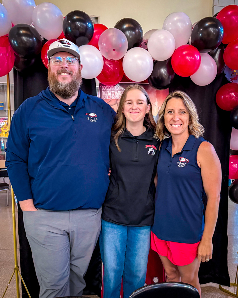A man and two women stand close, smiling, with a backdrop of colorful balloons and a black curtain.