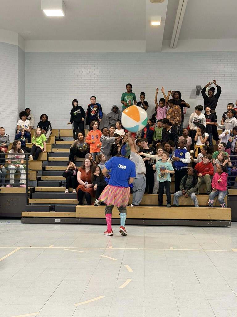 Students in the bleachers are reaching to hit a beach ball. 