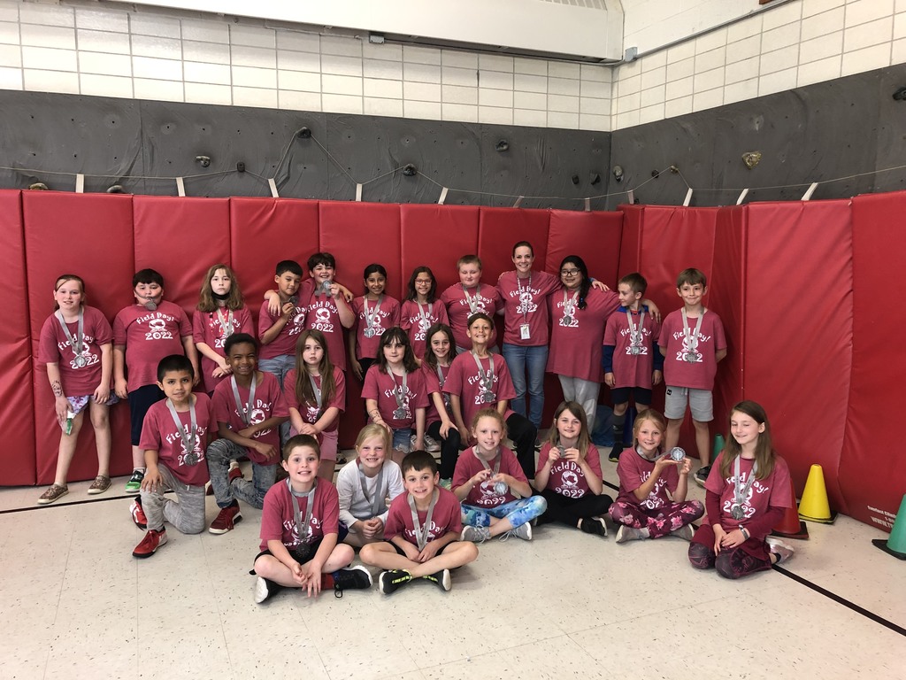 Mrs. Bohl's class is in front of the wall mats in the gym.  Some students are standing and some are sitting on the floor.