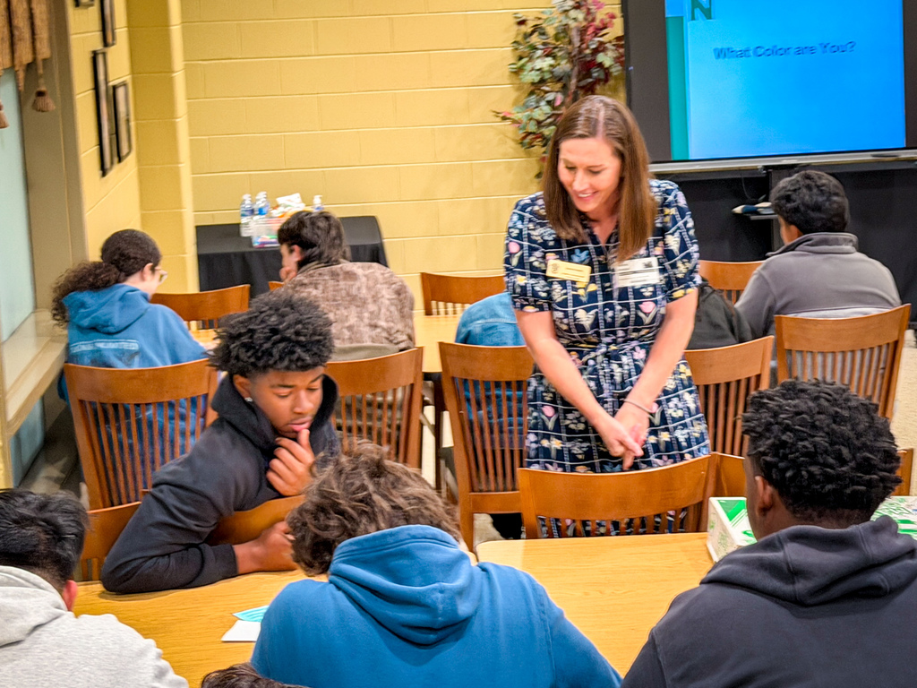 Woman teaching in classroom. Students in hoodies sitting at tables, listening. Bright yellow walls and projector screen in background.
