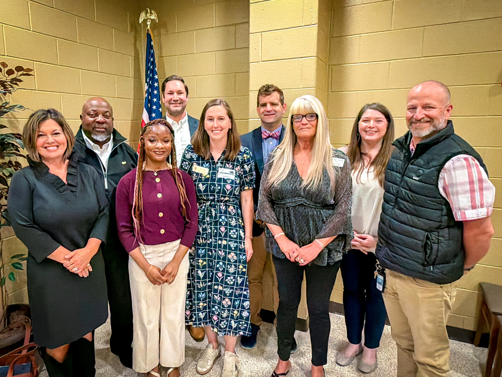 A group of people in formal attire stand together indoors. A man with a beard wears a vest, and a woman in a black dress holds a handbag.