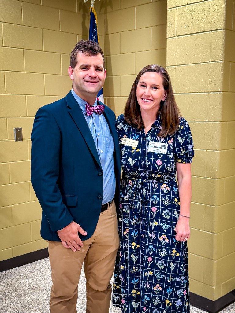 A man in a suit and a woman in a floral dress stand together in a room with yellow brick walls.