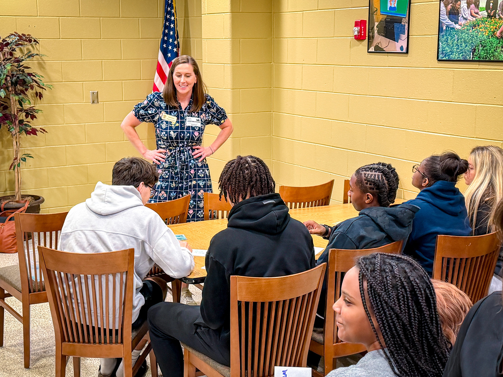 A woman stands in front of a group of people in a yellow room, behind her is a flag.