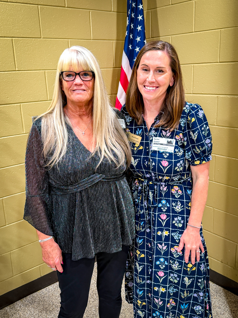 Two women stand close to each other in front of a yellow wall with a flag.