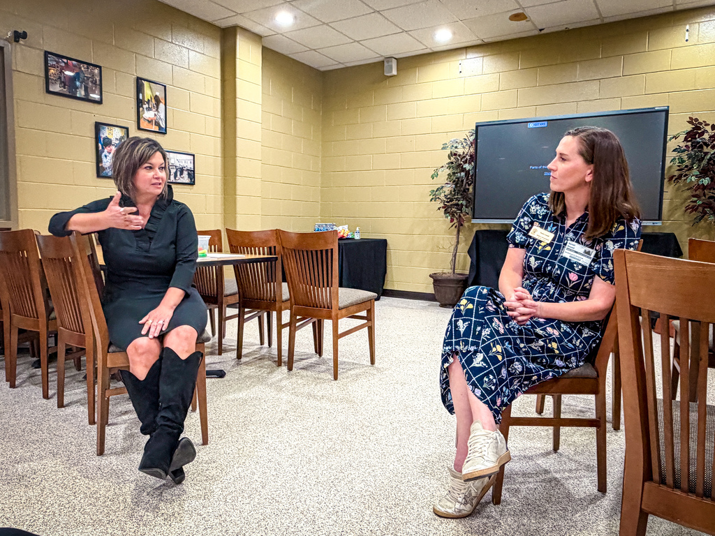 Two women sit in chairs inside a meeting room, one gesturing. Behind them, a table, chairs, and a large screen. Framed photos on walls.