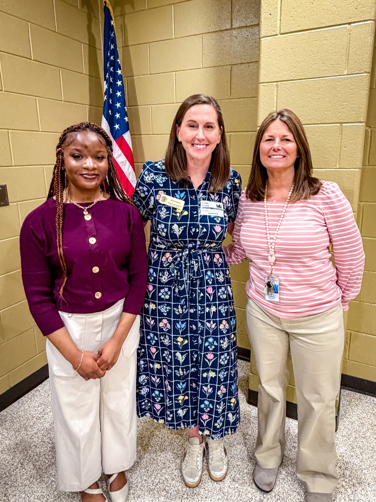 Three women standing together. One has a name tag. One wears a floral dress. Behind them is a flag.