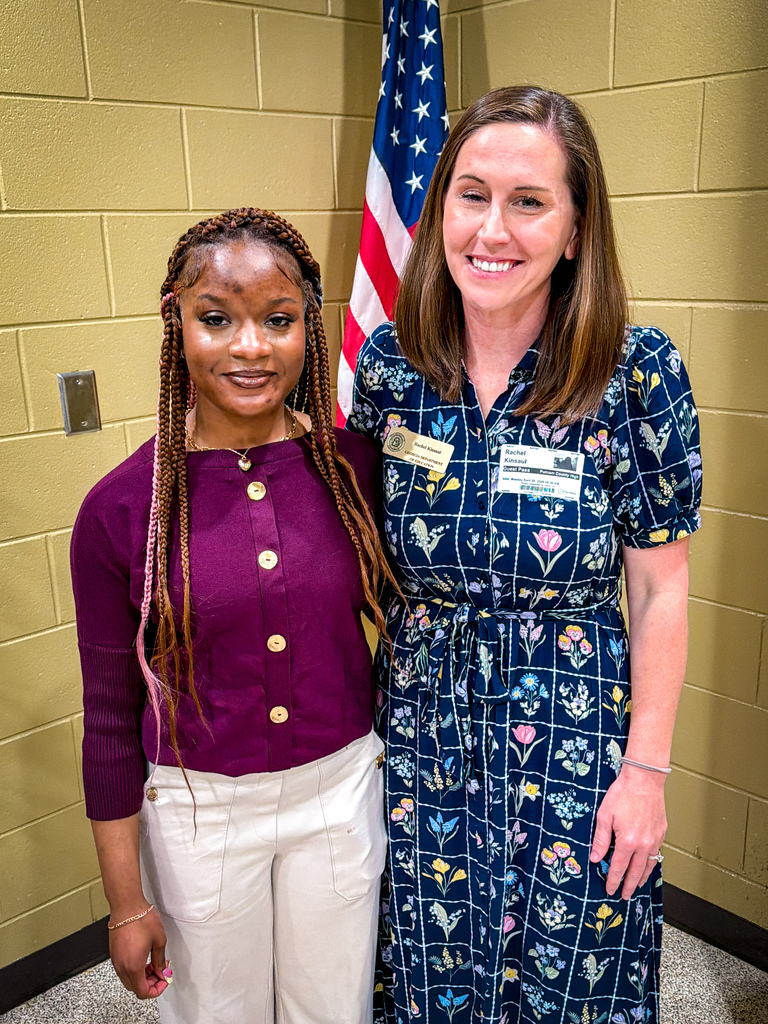 Two women stand together, one with braided hair and the other with a flower-patterned dress. An American flag is behind them.
