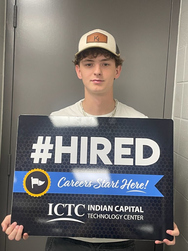 A young man wearing a white cap and necklace holds a sign that reads "#HIRED Careers Start Here!" with a blue ribbon.
