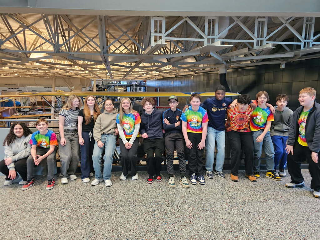Junior High students sit together outside the Steven F. Udvar-Hazy Center, part of the Smithsonian National Air and Space Museum. With their arms around one another, they smile warmly, conveying friendship, connection, and shared excitement during their visit.