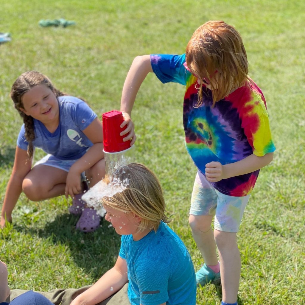 student in dumps water onto other student on field outside while another watches