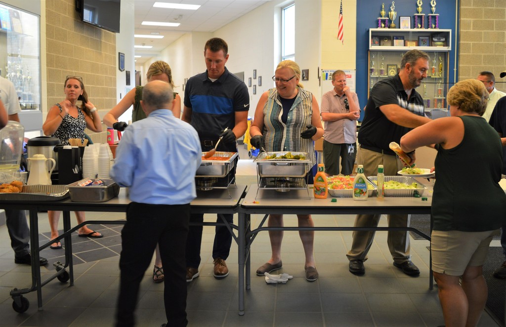 Four people serve food at a table as two people go through the line