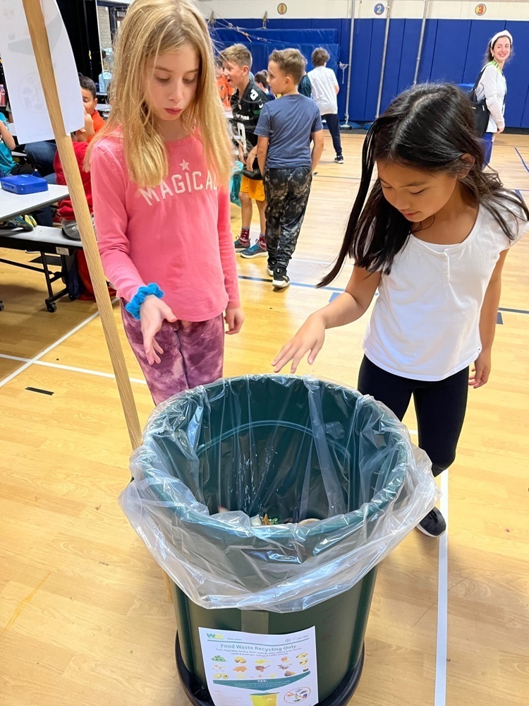 Two students places leftover food into the green barrel.