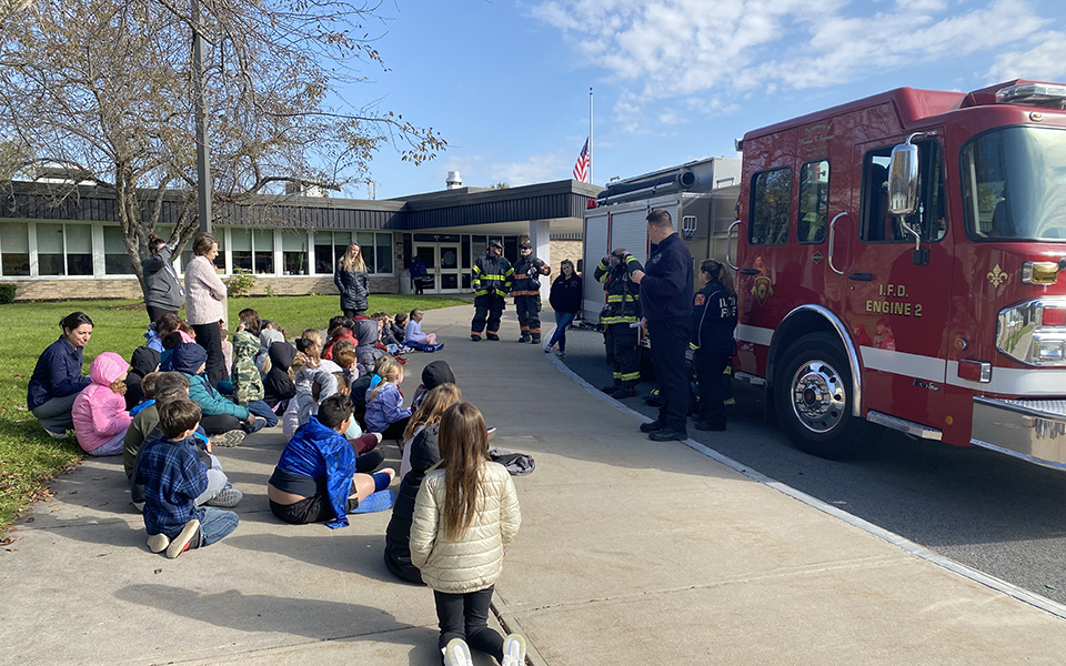 fire truck, school, students seated on sidewalk, fireman, sky, grass