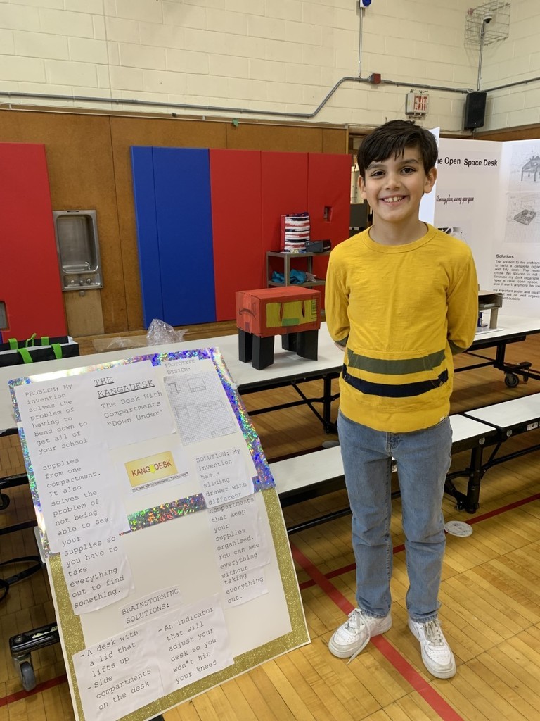 4th grade students in yellow shirt and jeans with poster board and protype of student desk in school gymnasium