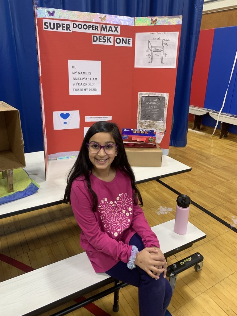 4th grade student in pink shirt sitting on bench with red poster board presentation in school gymnasium