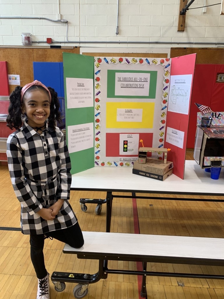 4th grade students in black and white checkered shirt with green, yellow, red and white poster board presentation in school gymnasium