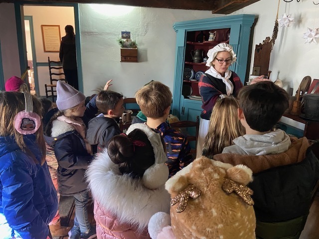 second grade class standing in a room at the Miller Cory House museum, listening to a woman speak
