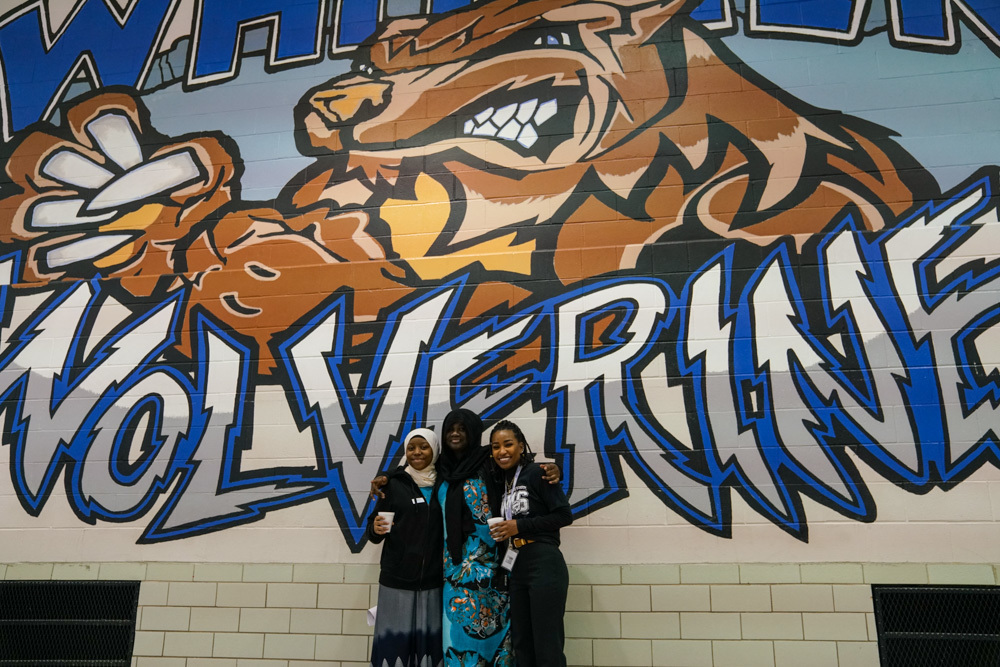 three women stand in front of a Whittier Middle School mural of a wolverine in the gym