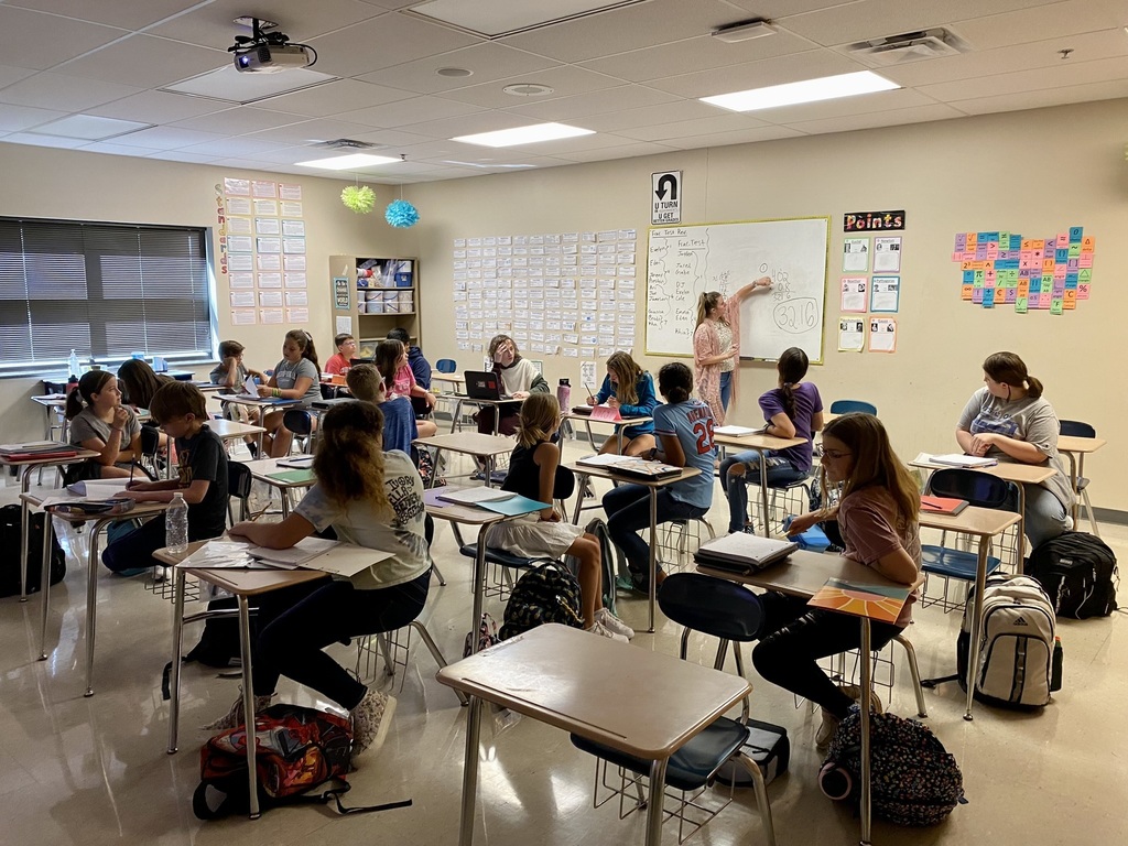 seventh grade students sit at desks in a classroom working on math. Mrs. Kaighin stands up front at a whiteboard explaining a problem