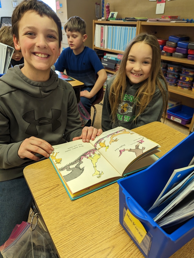 a 4th grade student reads a book with a 1st grade student on the floor