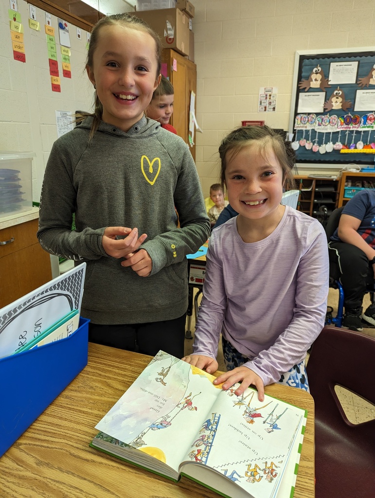 a 4th grade student reads a book with a 1st grade student on the floor
