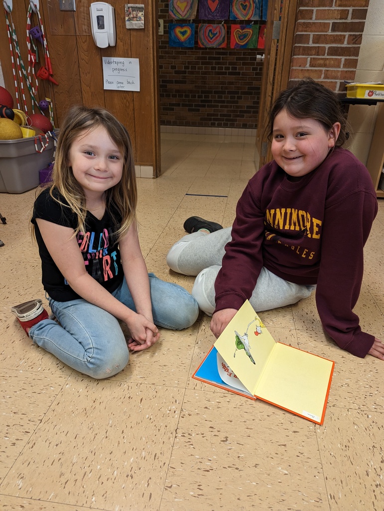 a 4th grade student reads a book with a 1st grade student on the floor