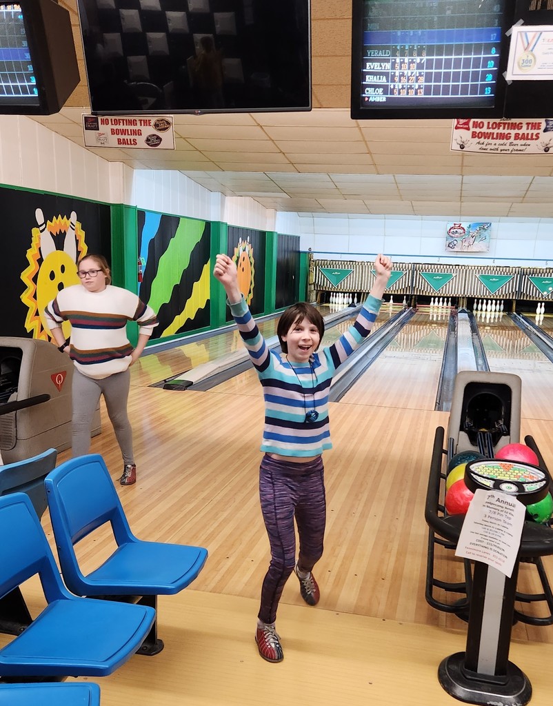 1 student excitingly stands on the bowling lane with arms in the air as another student also finishes throwing her ball