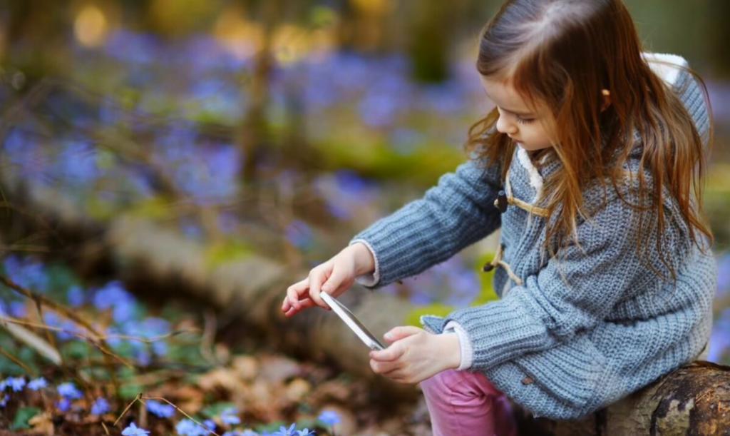 Little girl with long brown hair, wearing a blue sweater and pink pants is holding a cell phone outside, taking a photo of something on the ground.