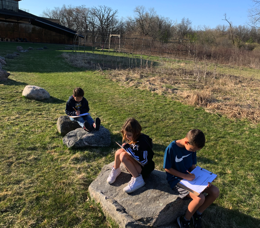 3 students working on homework outside, as they sit on large grandfather stones.