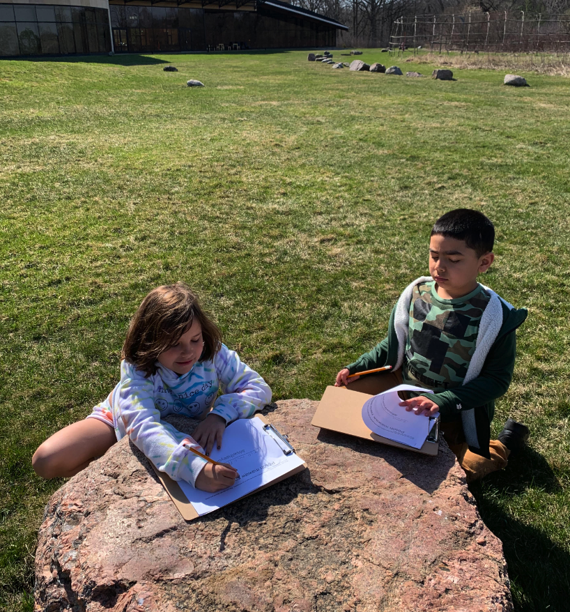 Two children outside, working on homework, using a large grandfather stone as their desk.
