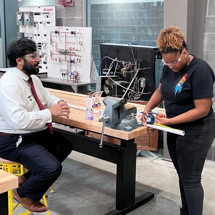 female student, putting something on a workbench, adult next to her talking to her