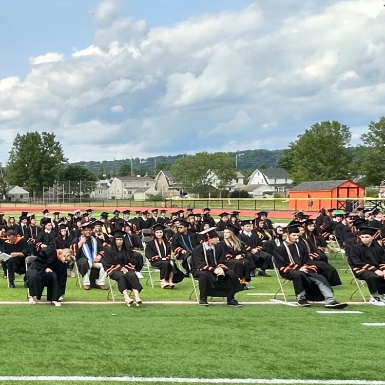 students sitting on a football field in black and orange graduation gowns 