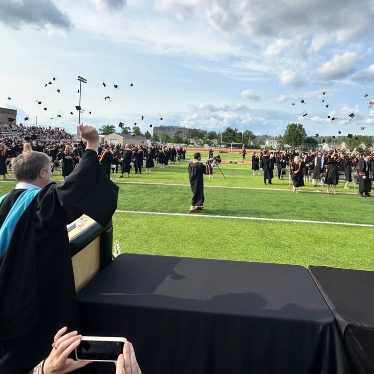 students on a football field in black and orange graduation gowns throwing their caps