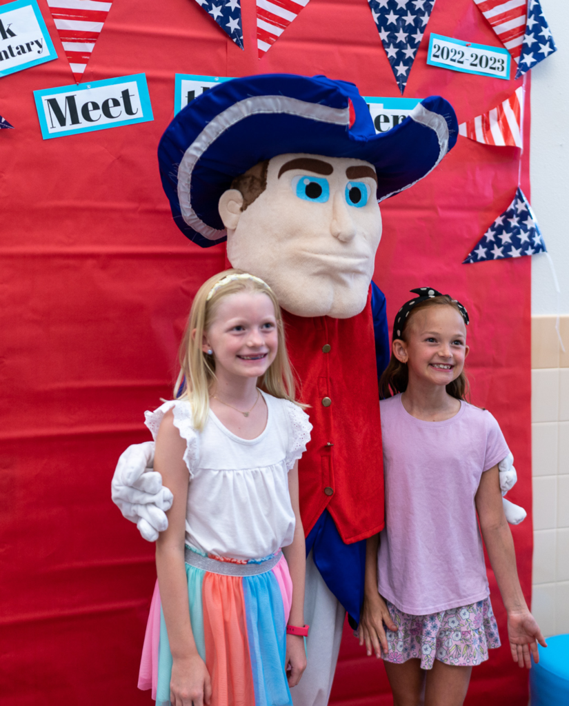 Caucasian girls in white and pink outfits posing for a photo with a patriot mascot in front of a red photo station.