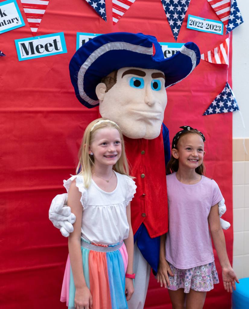 Caucasian girls in white and pink outfits posing for a photo with a patriot mascot in front of a red photo station. 