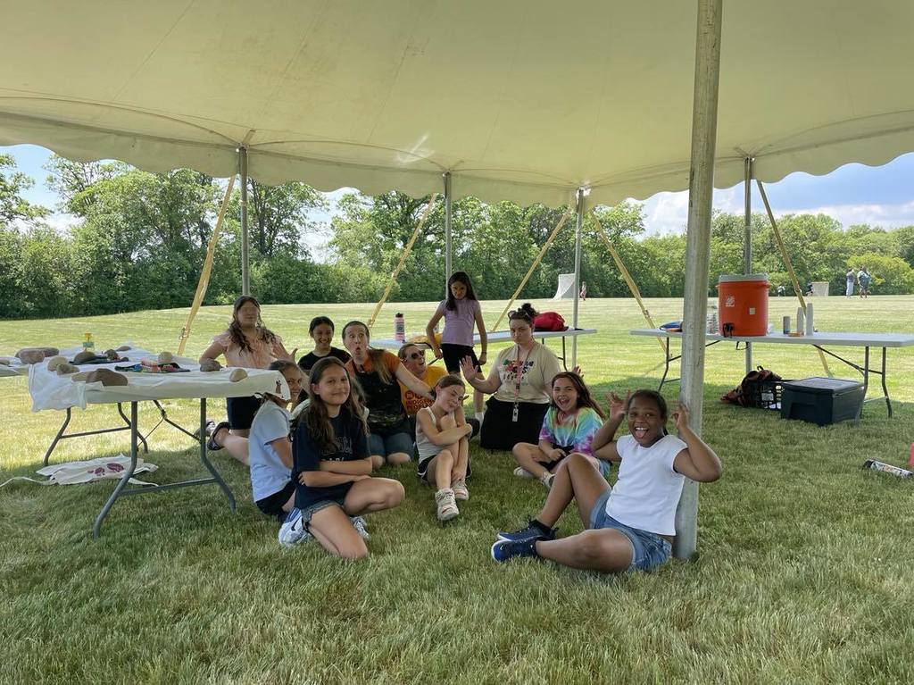Students sitting on the grass under the shade of a white tent.