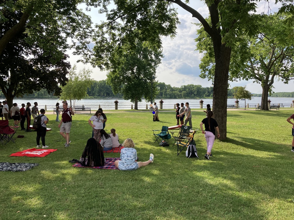 Park-like setting with many students walking around and sitting on blankets on the grass. Water, trees and blue sky are in this photo.