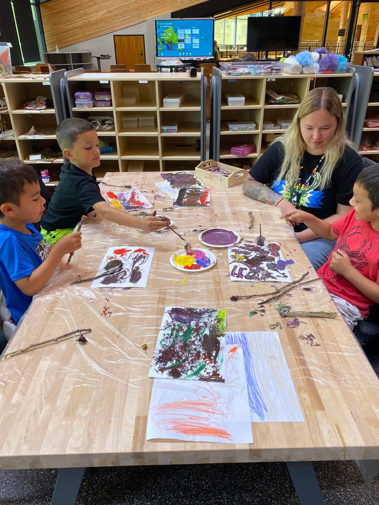 Three students painting on a table; one teacher sitting with them.
