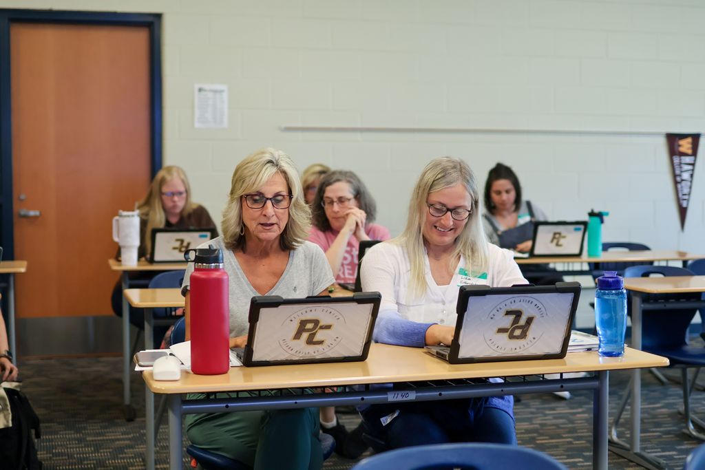 two women work on computers at a desk in a classroom