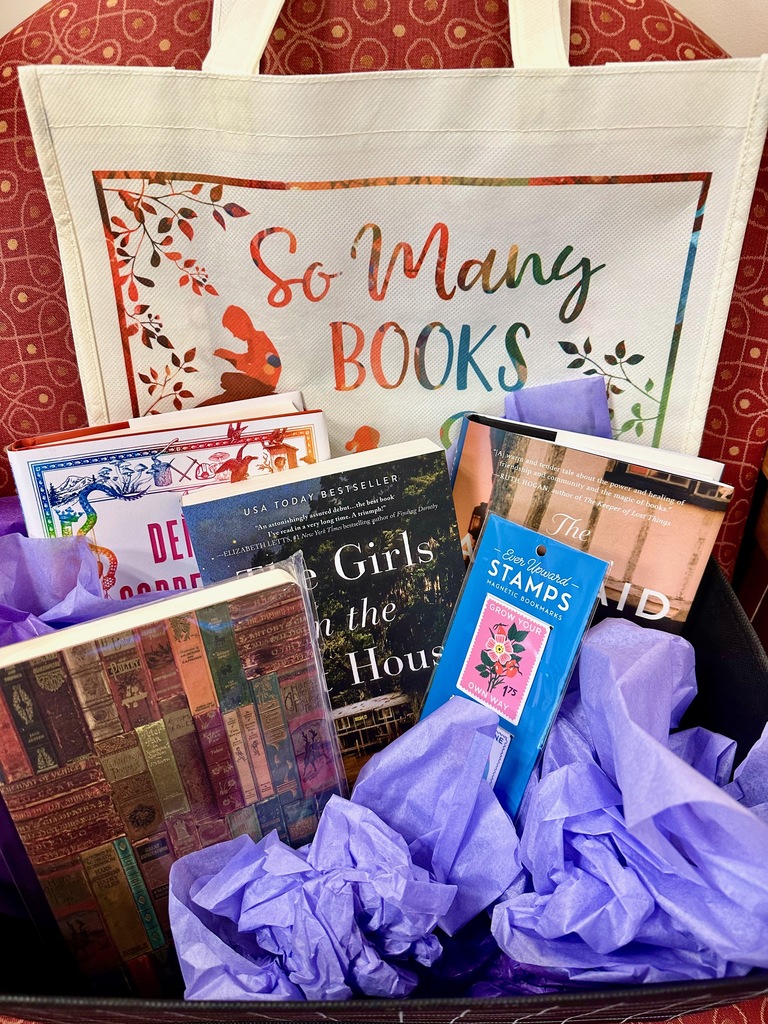 A library raffle basket includes a canvas bag, a book of stamps, a notebook, three books, and purple crepe paper